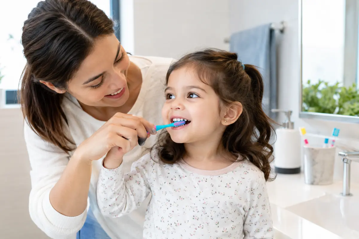 Parent helping a young child brush with fluoride toothpaste in a bright bathroom